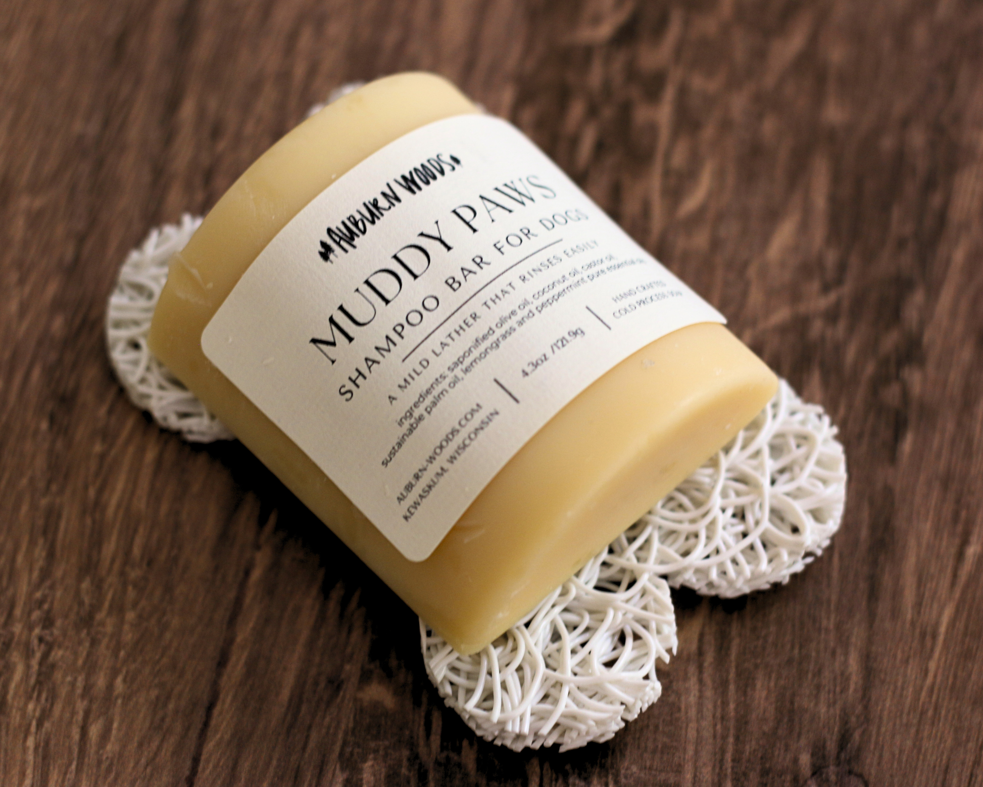 A bar of shampoo wrapped in a paper label reading 'Muddy Paws Shampoo Bar for Dogs' by #PupPupLover, resting on a white textured doily over a dark wooden surface.