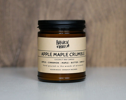 An amber glass jar candle with a black screw-top lid and a cream paper label reading Auburn Woods Apple Maple Crumble, set against a neutral wood and light gray background.
