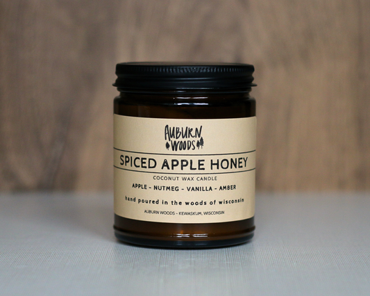 A jar candle labeled Auburn Woods Spiced Apple Honey, featuring a dark amber glass jar, cream-colored label, and black metal lid, set against a neutral wood and light gray background.