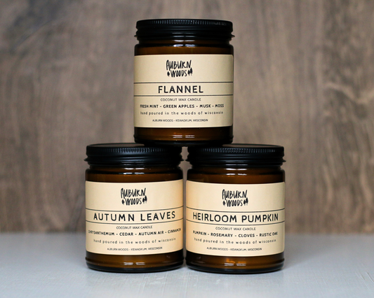 Three amber glass jar candles with black lids and cream labels from Harbourn Woods, stacked in a pyramid. The top candle is labeled 'FLANNEL', and the bottom two are labeled 'AUTUMN LEAVES' and 'HEIRLOOM PUMPKIN'.