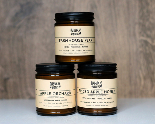 Three amber glass jar candles with black lids and cream labels from Auburn Woods, stacked in a pyramid on a light surface against a gray wood-textured background. The labels read Farmhouse Pear, Apple Orchard, and Spiced Apple Honey.